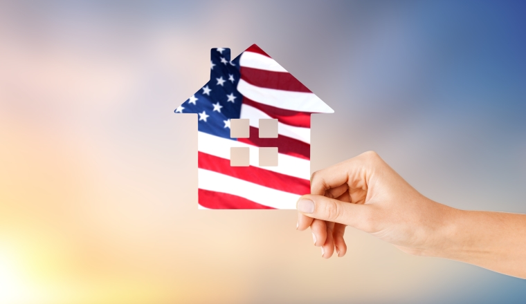patriotism, home and citizenship concept - hand holding paper house in colors of american flag over evening sky background