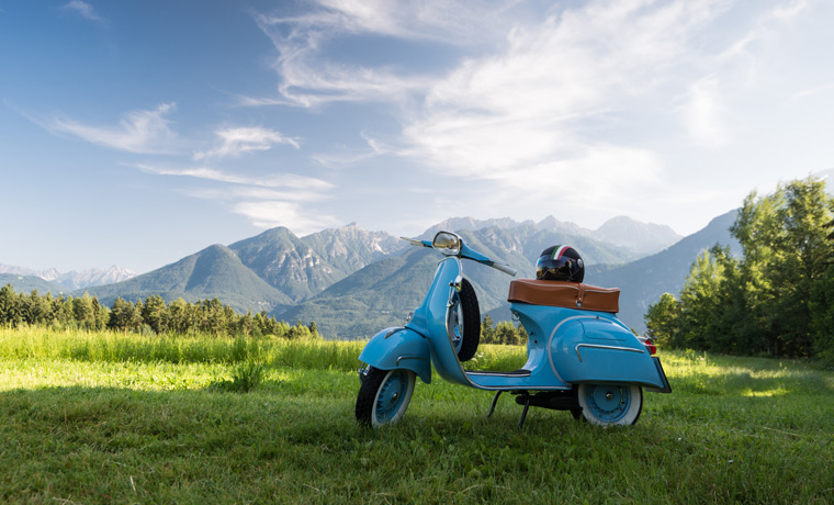 Blaue Vespa vor Bergkulisse auf Feld.