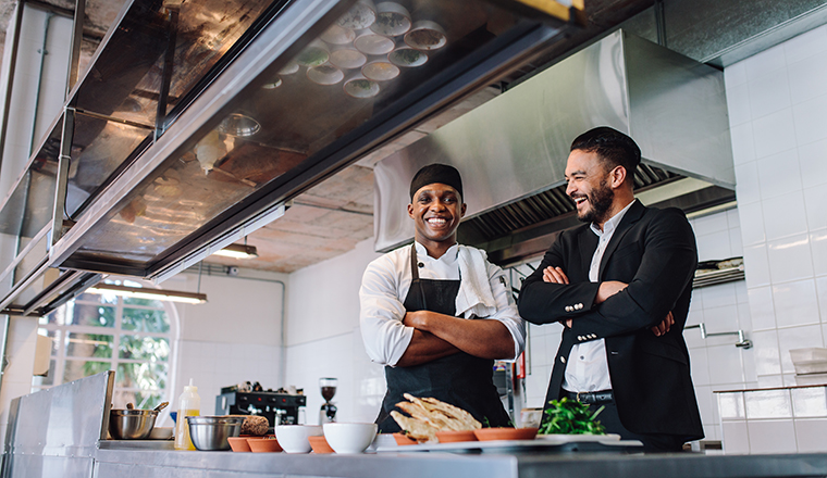 Smiling restaurant owner and chef standing in kitchen. Businessman with professional cook standing together and laughing.