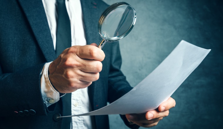 Tax inspector investigating financial documents through magnifying glass, forensic accounting or financial forensics, inspecting offshore company financial papers, documents and reports.