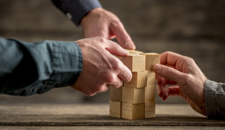 Hands of three people building a tower of wood blocks on a table, teamwork concept.