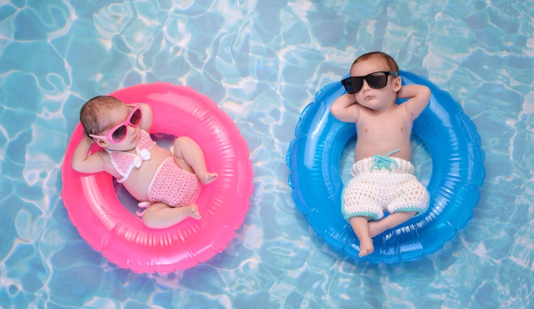 Two month old twin baby sister and brother sleeping on tiny, inflatable, pink and blue swim rings. They are wearing crocheted swimsuits and sunglasses.
