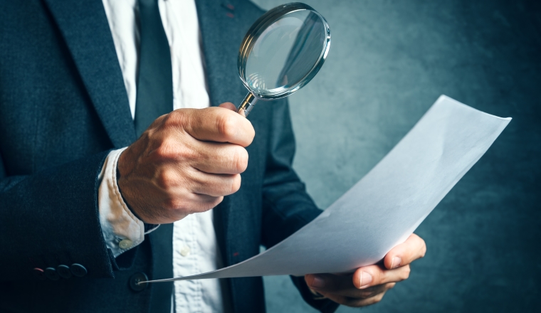 Tax inspector investigating financial documents through magnifying glass, forensic accounting or financial forensics, inspecting offshore company financial papers, documents and reports.