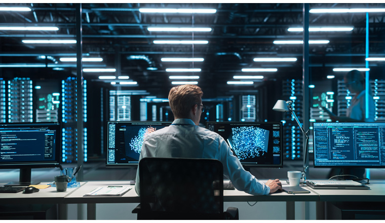 Software Developer Works with AI Analysis on his Computer with Two Monitors. Developer Coding while Looking at the Display of Computer. Modern Evening Dark Office. Back view.