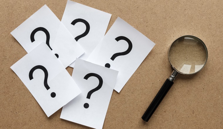 Question marks printed on white cards lying on a wooden background with a magnifying glass in a conceptual image