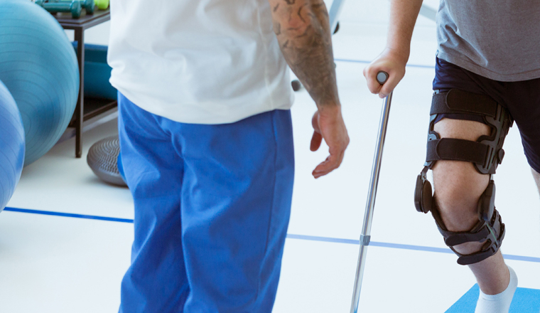 Man after car accident in an orthosis and on crutches learning to walk in the clinic, helpful therapist near him