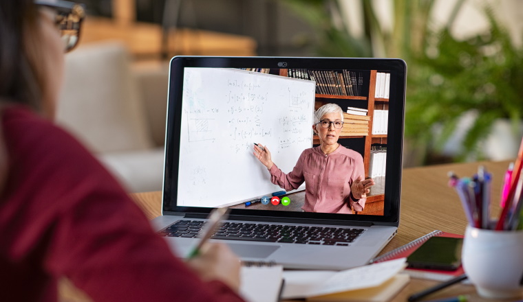 Young student watching lesson online and studying from home. Young woman taking notes while looking at computer screen following professor doing math on video call. Latin girl student studying from home and watching teacher explaining math formula on video chat.