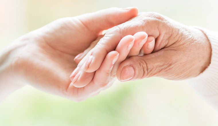 Close-up of tender gesture between two generations. Young woman holding hands with a senior lady. Blurred background. Panorama.