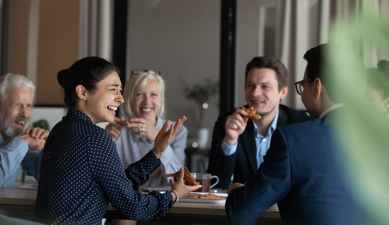 Excited diverse employees eating pizza during break in office together, happy Indian businesswoman laughing at funny joke, talking chatting with colleagues, having fun, sharing corporate lunch