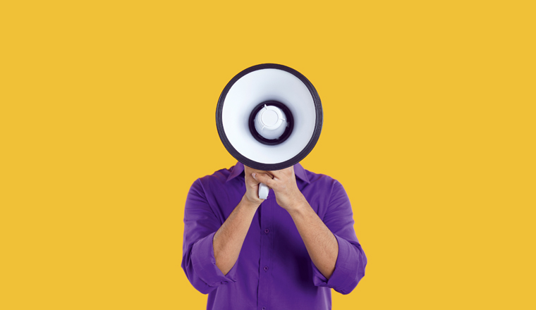 Man hiding his face behind loudspeaker makes loud announcement or advertisement on orange background. Unknown man in purple casual shirt holding bullhorn in front of his face. Advertisement concept.