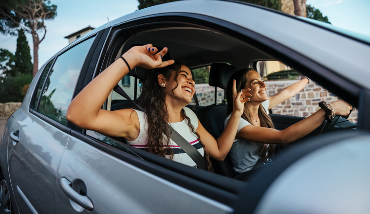 Two young women sing a song on the radio and dance in the car on a day trip in the summer - Best friends having fun together driving around the countryside - Smiling millennial in a relaxing moment