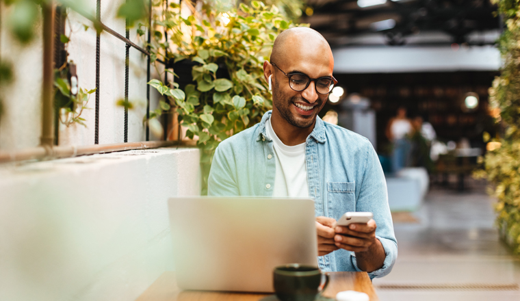 Man sits in a cafe, casually dressed and smiling while using his smartphone to play music from a streaming app. Happy young man relaxing with a laptop and a cup of coffee on the table nearby.