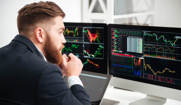 Back view of thoughtful bearded young businessman sitting and woking with graph and charts on computer