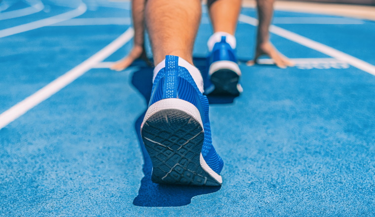 Sprinter fit man waiting for start of race on running tracks at outdoor stadium. Sport and fitness runner athlete on blue run track starting line with running shoes. Banner panorama.