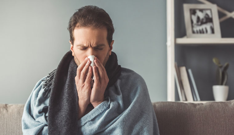 Handsome sick man is wiping nose while sitting on couch covered in plaid