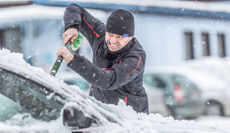 Male driver franatically scrapes ice from frozen windshields of his car parked outdoors.