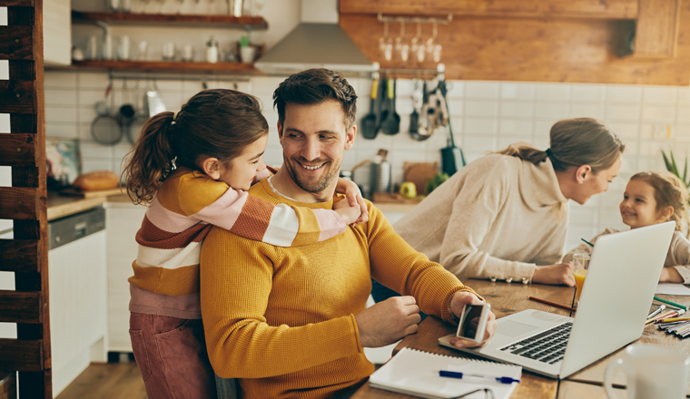 Happy little girl embracing her father and talking to him while he's working on a computer at home.