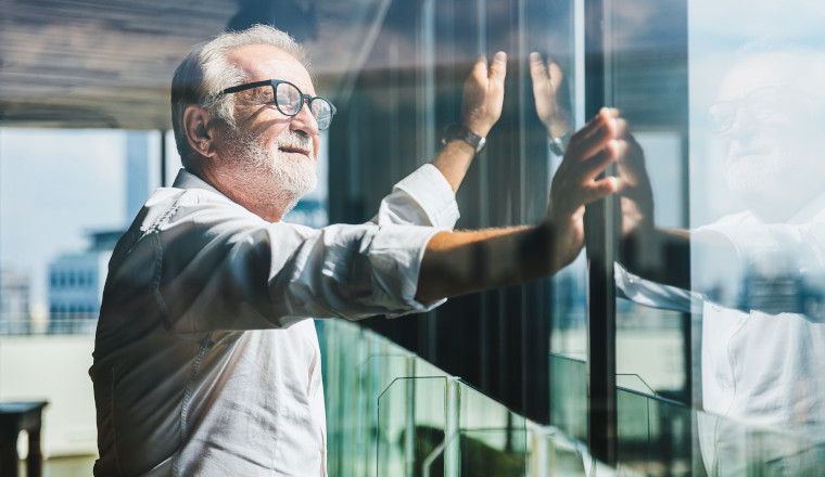 Retirement concept. Senior grey-haired businessman standing and looking to right hand at modern business lounge high up in an office tower.