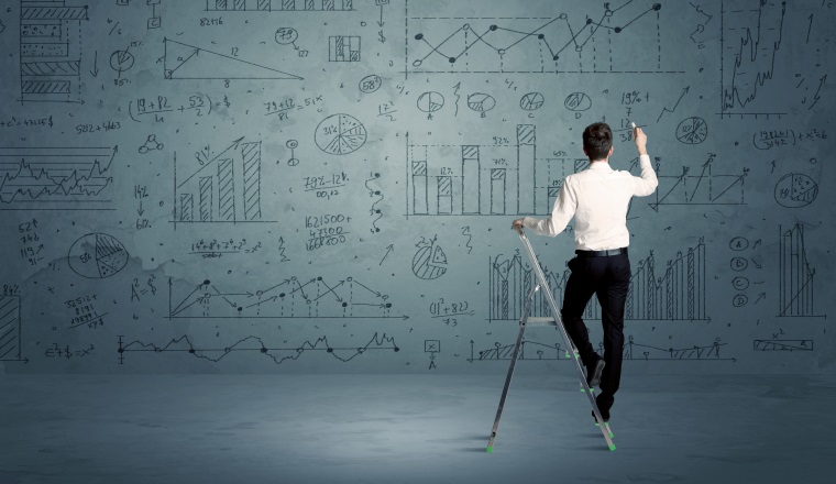 A businessman in modern stylish elegant suit standing on a small ladder and drawing pie and block charts on grey wall background with exponential progressing curves, lines, circles, angles, blocks, numbers