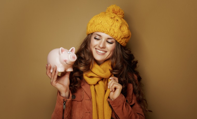Portrait of smiling elegant woman in yellow beret and scarf looking at piggy bank against brown background.