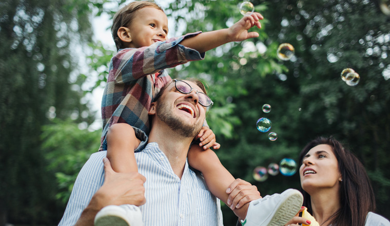 Happy young family playing with bubble wands with son sitting on fathers shoulders in park outdoors in summer; Shutterstock ID 711949186; purchase_order: Blueroom Photo Library Feb 2022; job: ; client: ; other: 