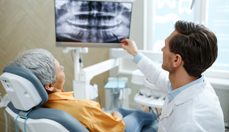 Back view of male dentist pointing at tooth X-ray image on screen during consultation in modern dental clinic