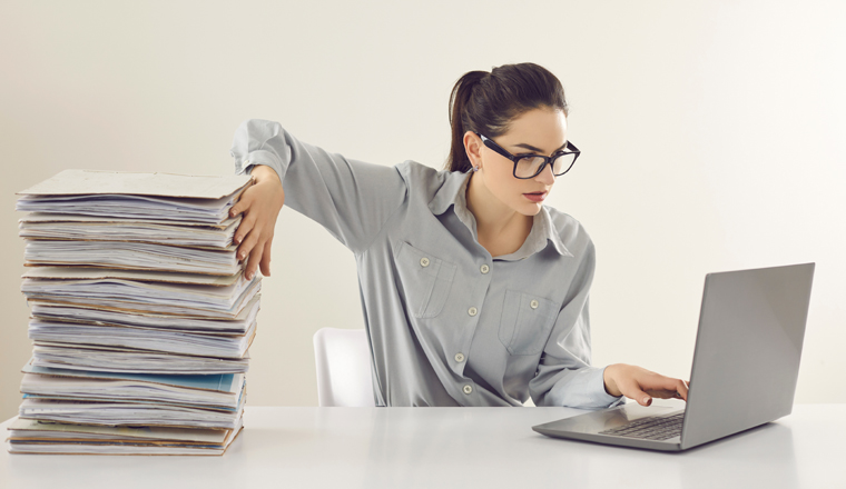 Young accountant working on laptop computer sitting at desk with pile of papers. Paperwork vs electronic documents. Storing files in digital database. Having quick convenient access to storage system