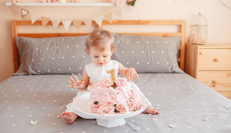 Portrait of cute adorable Caucasian blonde baby girl in white dress celebrating her first birthday. Home indoors cake smash first year concept