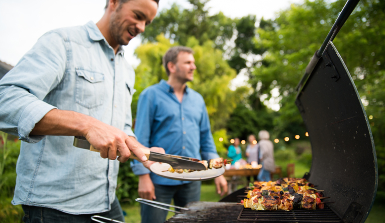 n a summer evening,  two men  in their forties prepares a barbecue for  friends gathered around a table in the garden