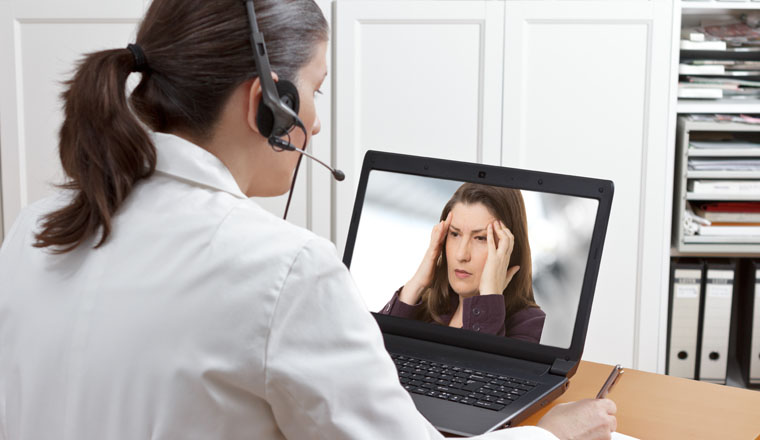 Doctor sitting at the desk of her office with headset and laptop, making a video call with a patient suffering from acute headache, telehealth or telemedicine concept