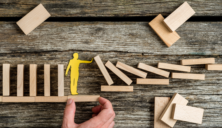 Conceptual close-up of the hand of a man offering support through a stable platform to a paper man stopping the collapse caused by domino effect.