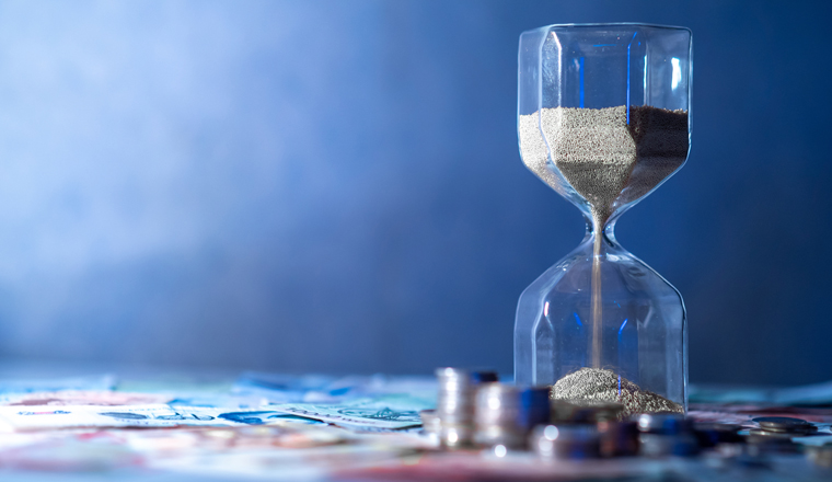 Sand running through the shape of hourglass on table with banknotes and coins of international currency. Time investment and retirement saving. Urgency countdown timer for business deadline concept