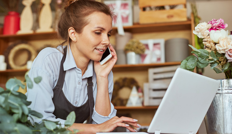 Portrait of florist in apron working at her own flower shop, using laptop and calling on the smart phone. She is leaning on wooden counter. She is arranging logistics, and delivery, taking orders