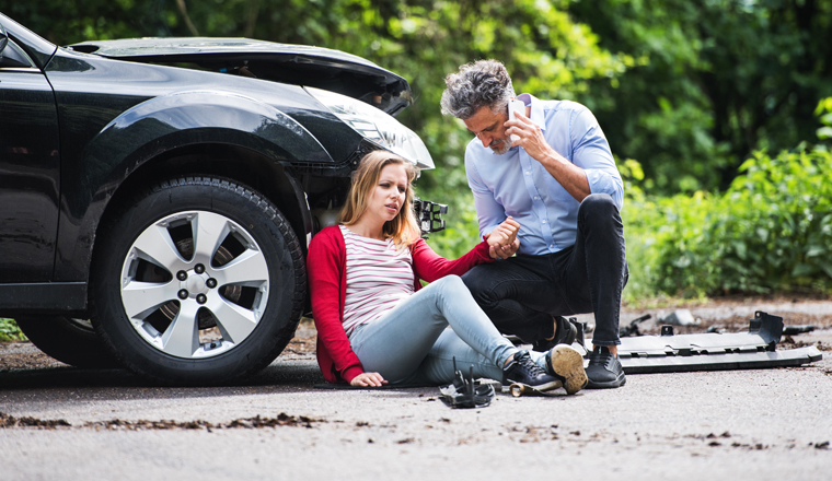 Young woman by the car after an accident and a man with smartphone, making a phone call.