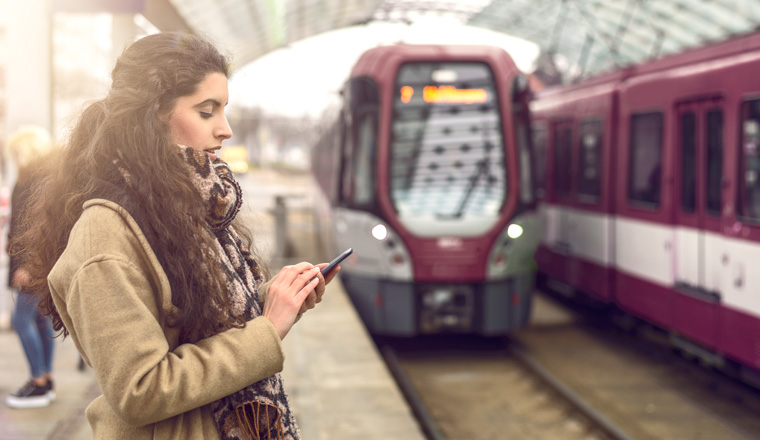 Smiling brunette young woman in coat, standing at station with approaching train while holding her mobile phone