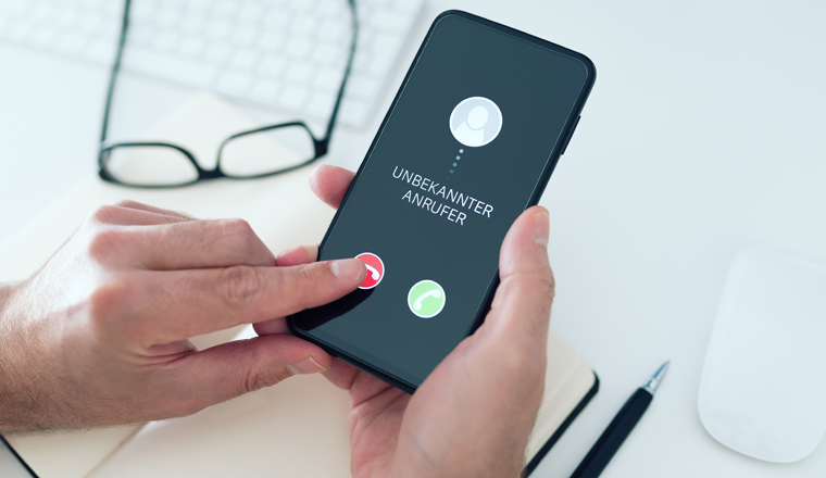 close-up view of person rejecting call from unknown number with text UNBEKANNTER ANRUFER, German for unknown caller, on smartphone, phone scam and phishing concept