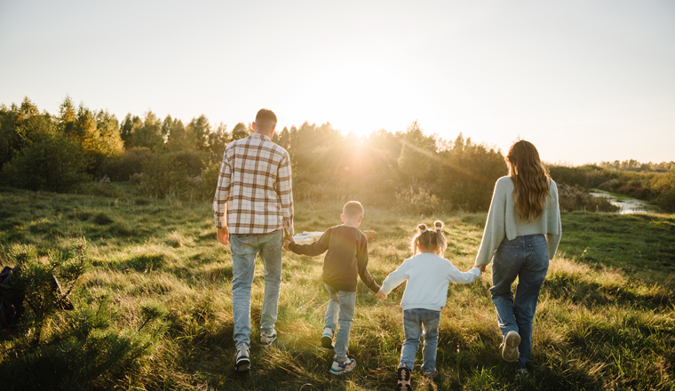 Mom, dad, daughter and son walk back in green grass in field. Happy young family with children spending time together, running outside, go in nature, on vacation, outdoors. Concept of family holiday.